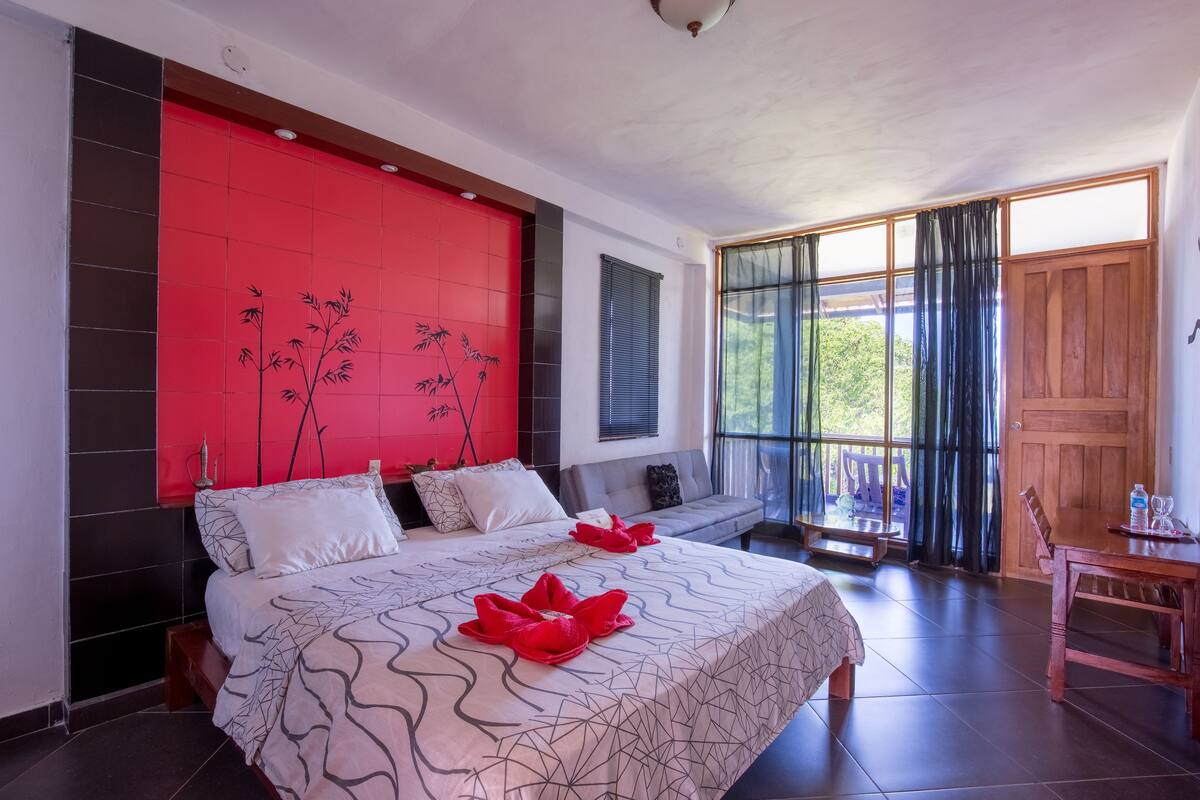 A modern hotel room with a large bed, a red accent wall, and a view of the outdoors.