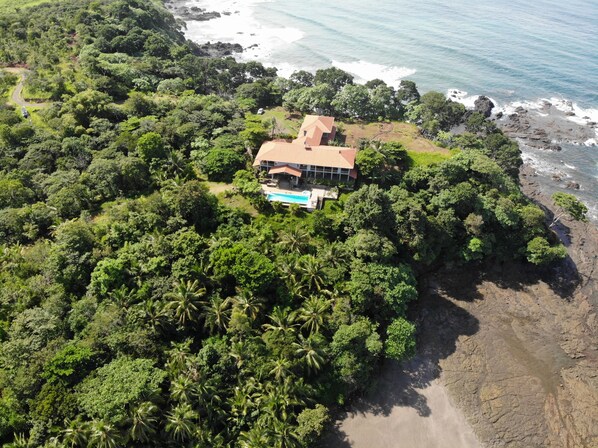An aerial view of a house surrounded by dense tropical vegetation, with a visible swimming pool and a rocky shoreline.
