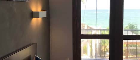 A hotel room with a bed, a nightstand, a painting on the wall, and a view of the ocean through large windows.