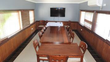 Un salón de conferencias con una larga mesa de madera, sillas, un televisor montado en la pared y ventanas con persianas.