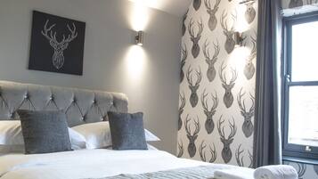 A bedroom with a tufted headboard, a bed with grey bedding, two pillows, a wall-mounted light, and a window with curtains.