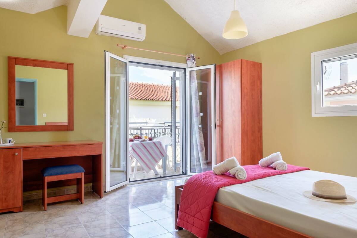 A bedroom with a bed, a desk, a chair, a mirror, and a sliding glass door leading to a balcony.