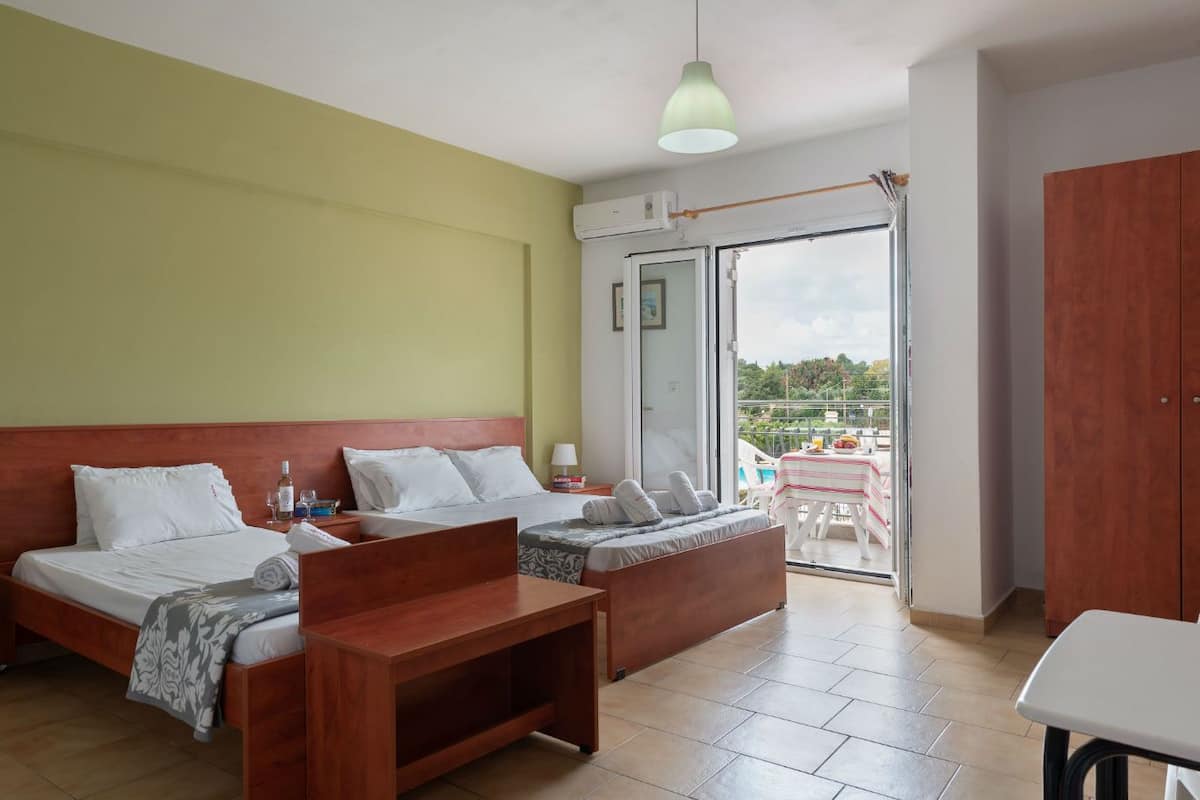 A hotel room with two beds, a wooden table, a balcony with a table and chairs, and a view of greenery.