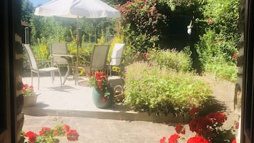 En vy genom en dörröppning in till en uteplats med utemöbler, en pergola med klätterväxter och krukväxter med röda blommor.