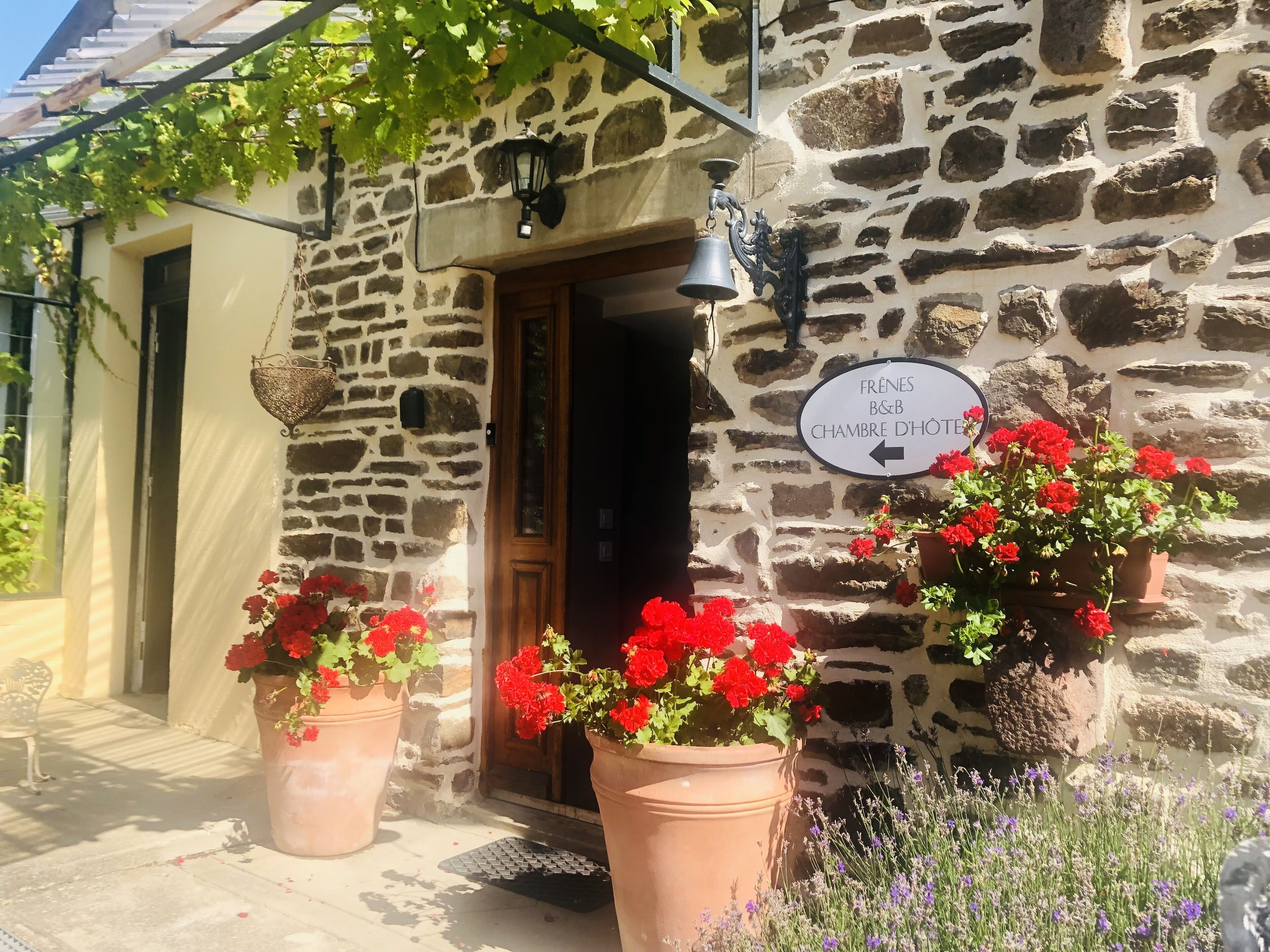 A stone building with a wooden door, potted red flowers, and a sign indicating a restaurant named "Frênes B&B Chambre d'Hôtellerie".