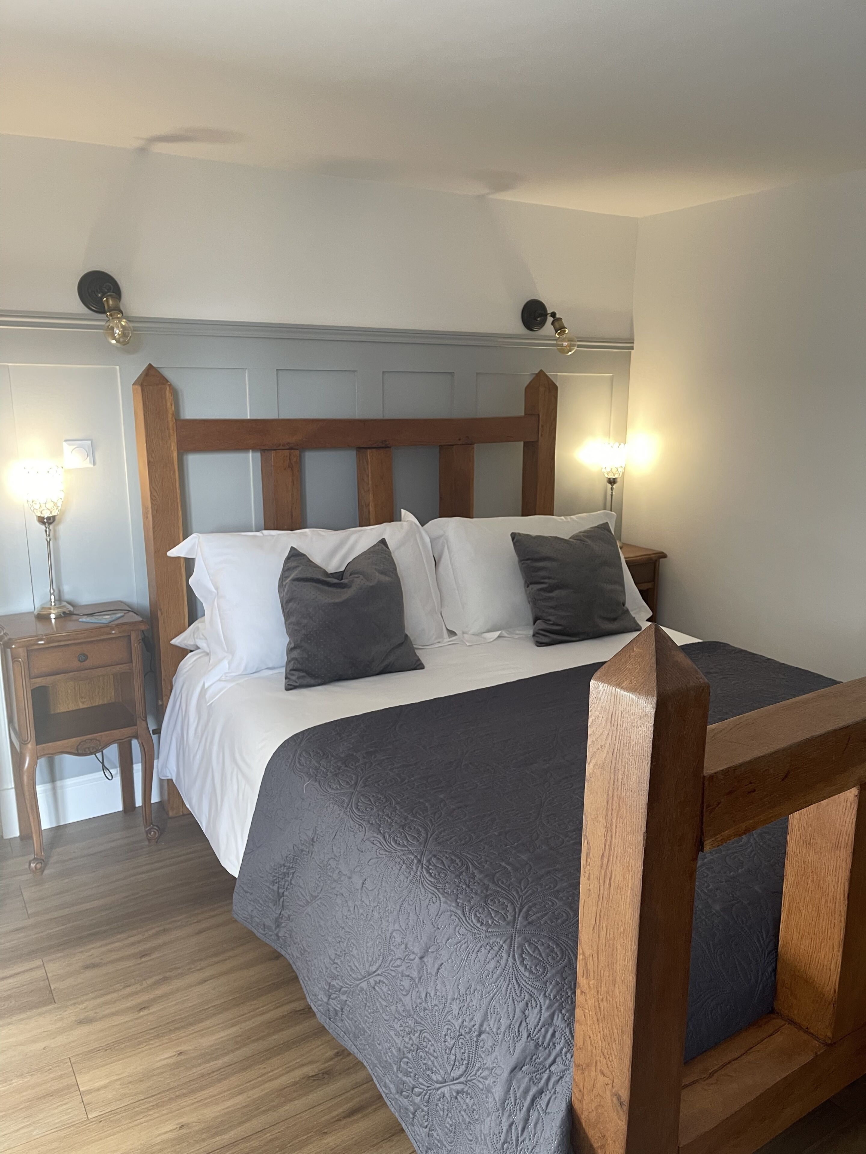 Ein ordentlich bezogenes Bett mit Holz-Kopfstück, ein Nachttisch mit Lampe und ein Beistelltisch mit Lampe.