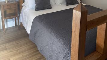 Ein ordentlich bezogenes Bett mit Holz-Kopfstück, ein Nachttisch mit Lampe und ein Beistelltisch mit Lampe.