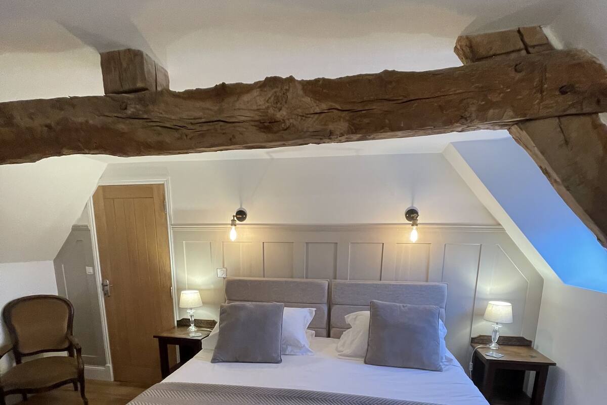 A bedroom with a sloped ceiling, a bed with grey bedding, two bedside lamps, and a wooden chair.