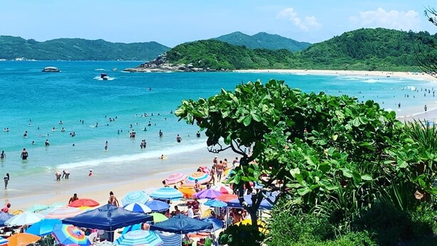 A beach with people swimming and sunbathing, surrounded by lush green hills. - Pousada Villa Maciel (Bombinhas)