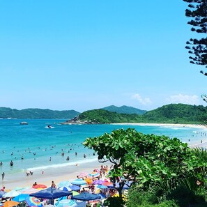 A beach with people swimming and sunbathing, surrounded by lush green hills. - Pousada Villa Maciel (Bombinhas)