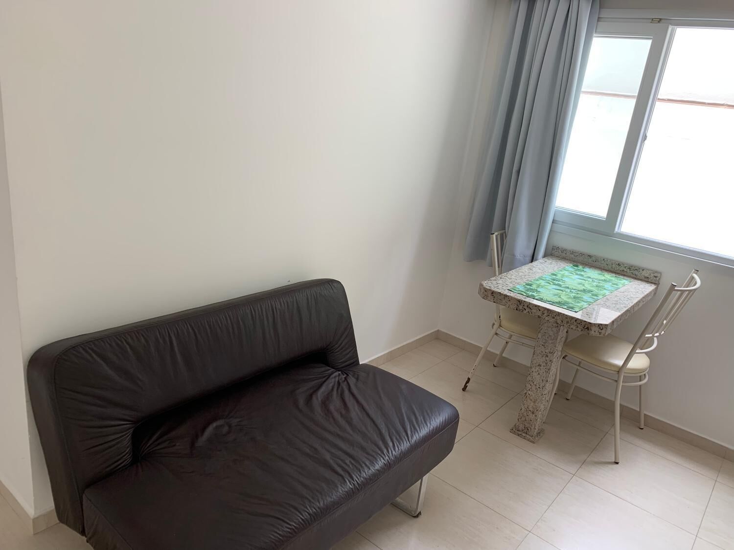 Una habitación con un sofá de cuero marrón oscuro, una mesita con mantel estampado verde y una ventana con cortinas azul claro.