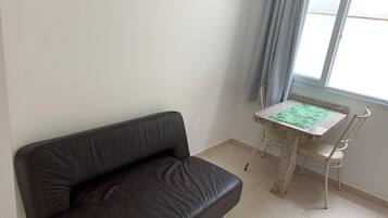 Una habitación con un sofá de cuero marrón oscuro, una mesita con mantel estampado verde y una ventana con cortinas azul claro.