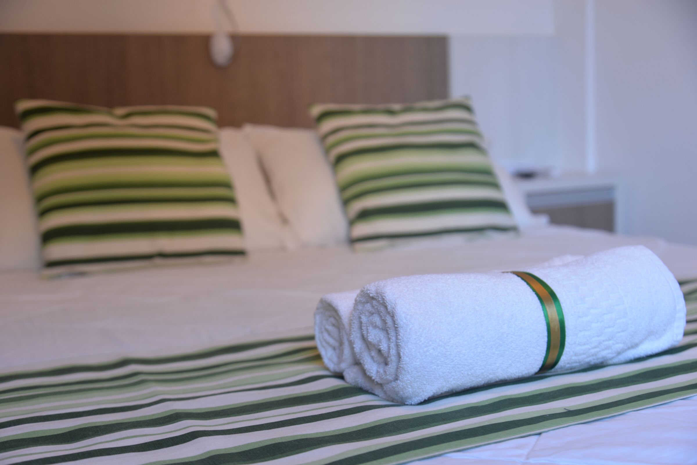 A neatly made bed with green and white striped bedding, a rolled white towel with a green band, and matching green and white striped pillows.