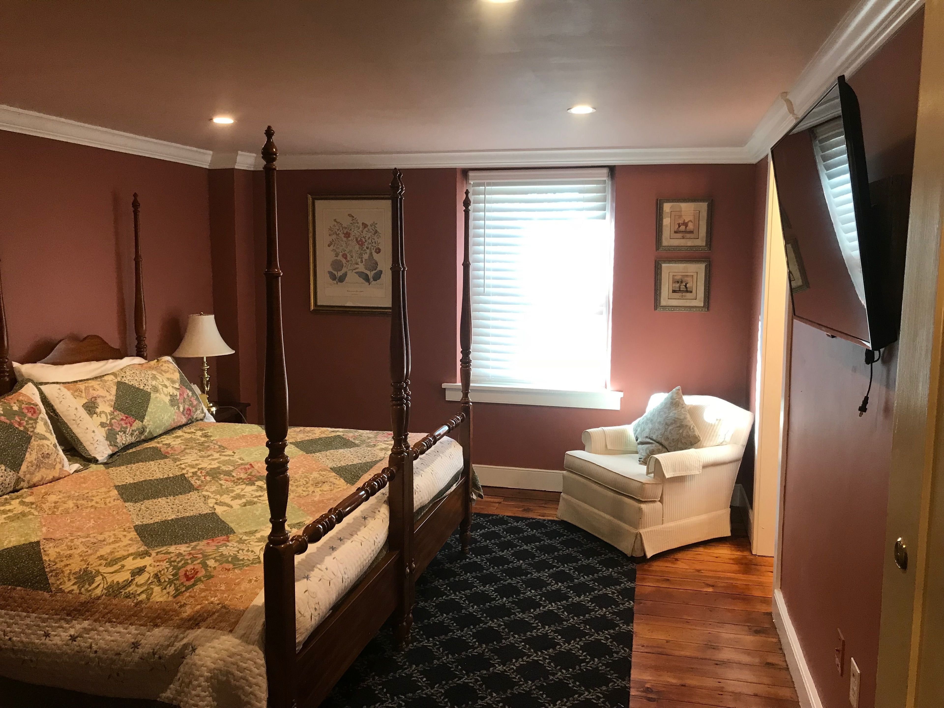 A bedroom with a four-poster bed, a chair, a window with blinds, and framed pictures on the wall.