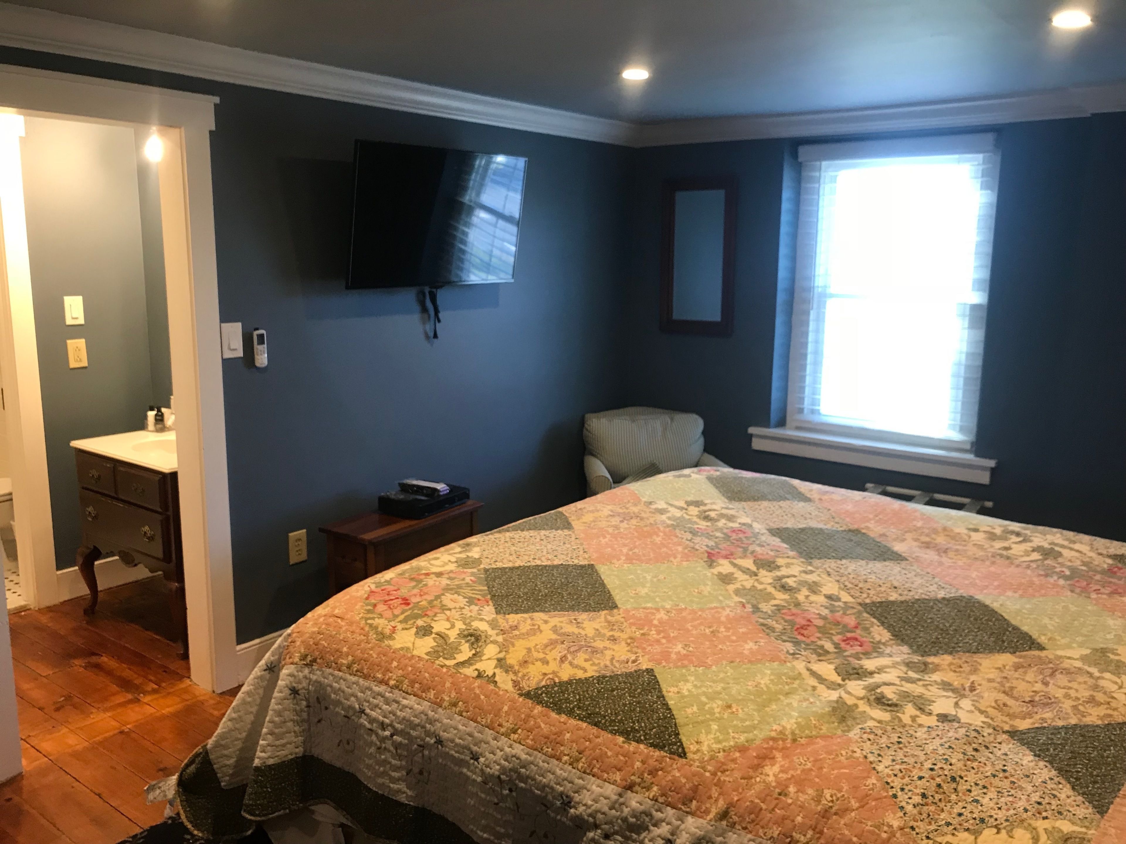 Quarto com cama grande, televisão na parede, janela com persianas e uma pequena mesa de cabeceira.