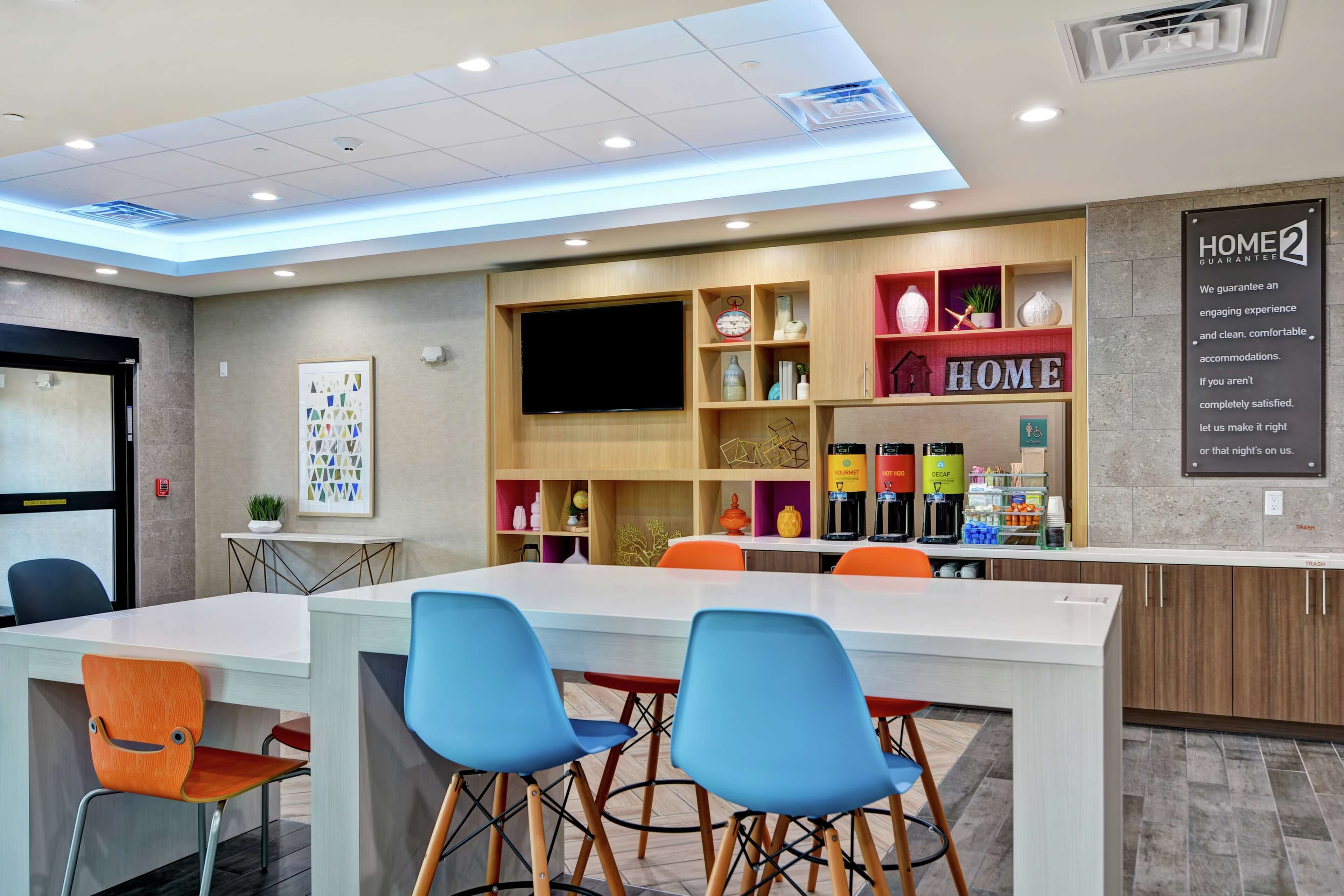 Een moderne hotel lobby met een receptiebalie, een grote flatscreen-tv en een bord waarop 'HOME' staat.