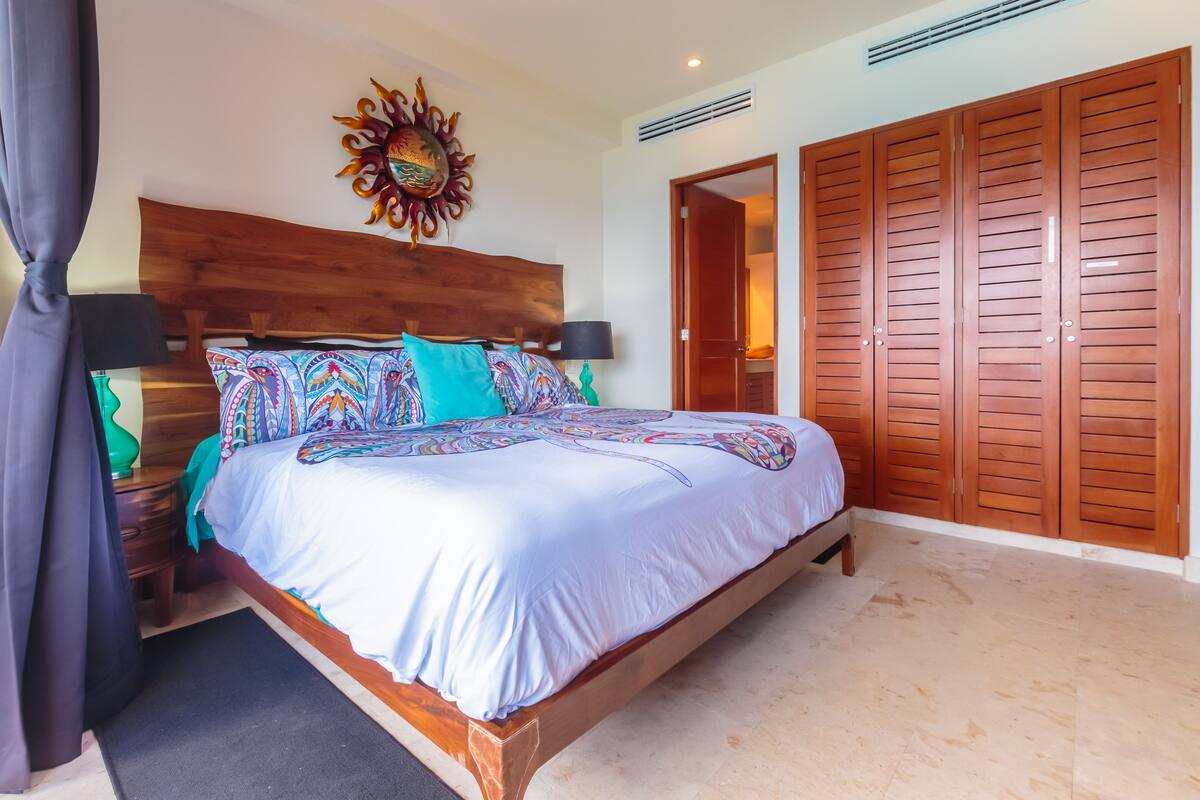 A bedroom with a wooden bed, patterned pillows, a decorative wall piece, and a wardrobe.