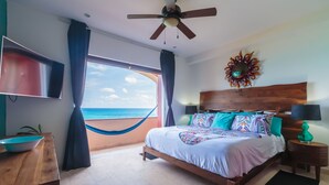 A bedroom with a bed, a ceiling fan, a TV, a wooden headboard, a nightstand, a lamp, and a view of the ocean.