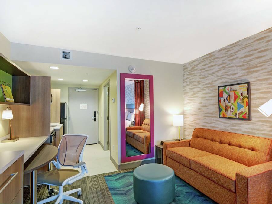 A hotel room with a desk, chair, orange sofa, round ottoman, and a mirror with a pink frame.