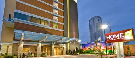A hotel entrance with a covered walkway, a large sign for 'Home 2 Suites by Hilton', and a cityscape view in the background.