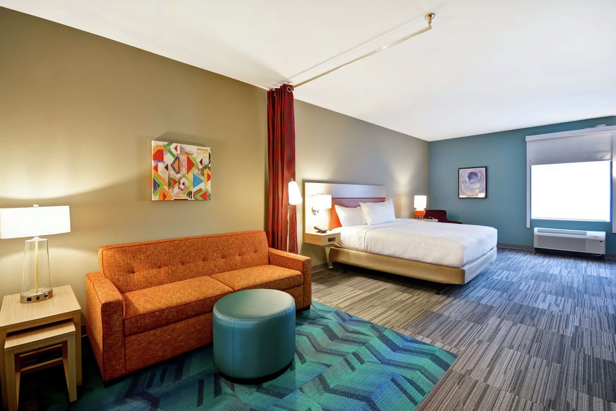Habitación de hotel con cama, sofá, una mesita, lámpara, alfombra colorida y ventana con cortinas.