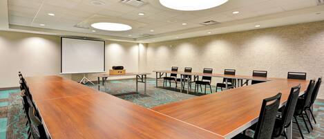 A conference room with a long wooden table, black chairs, a projector screen, and a brick-patterned wall.