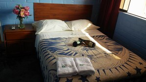 Een net opgemaakt bed met een beddengoed met een patroon, twee opgevouwen handdoeken, een fles en een afstandsbediening op een nachtkastje naast het bed.