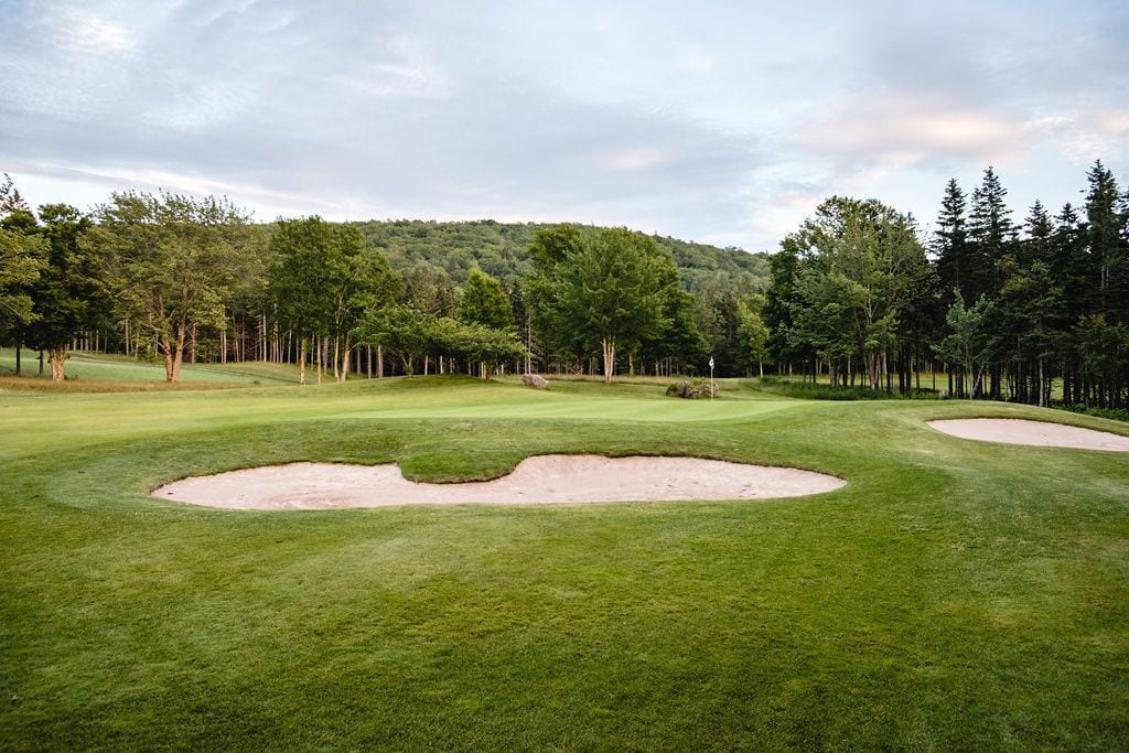 A golf course with sand bunkers, a well-maintained green, and a forested hill in the background.