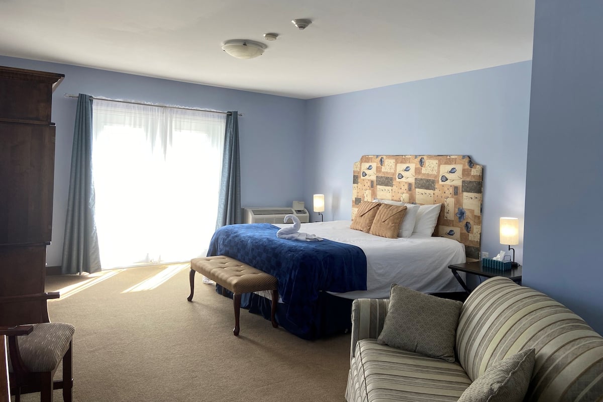 Ein Hotelzimmer mit einem Bett, einer Bank, einem Sofa, einem Schreibtisch mit Lampe und einem fenster mit Vorhängen.