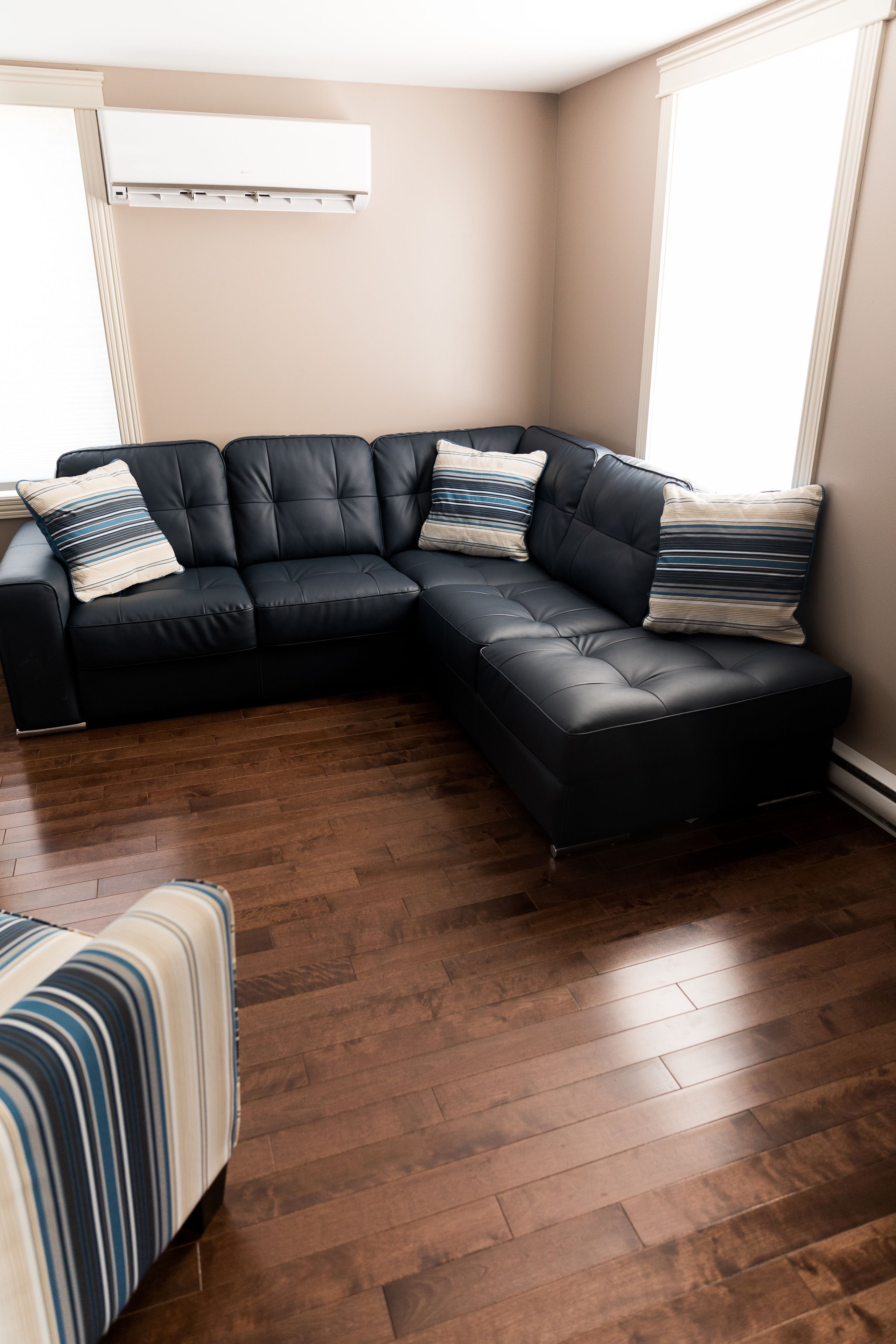 A corner living room with a black leather sectional sofa, striped pillows, and a wooden floor.