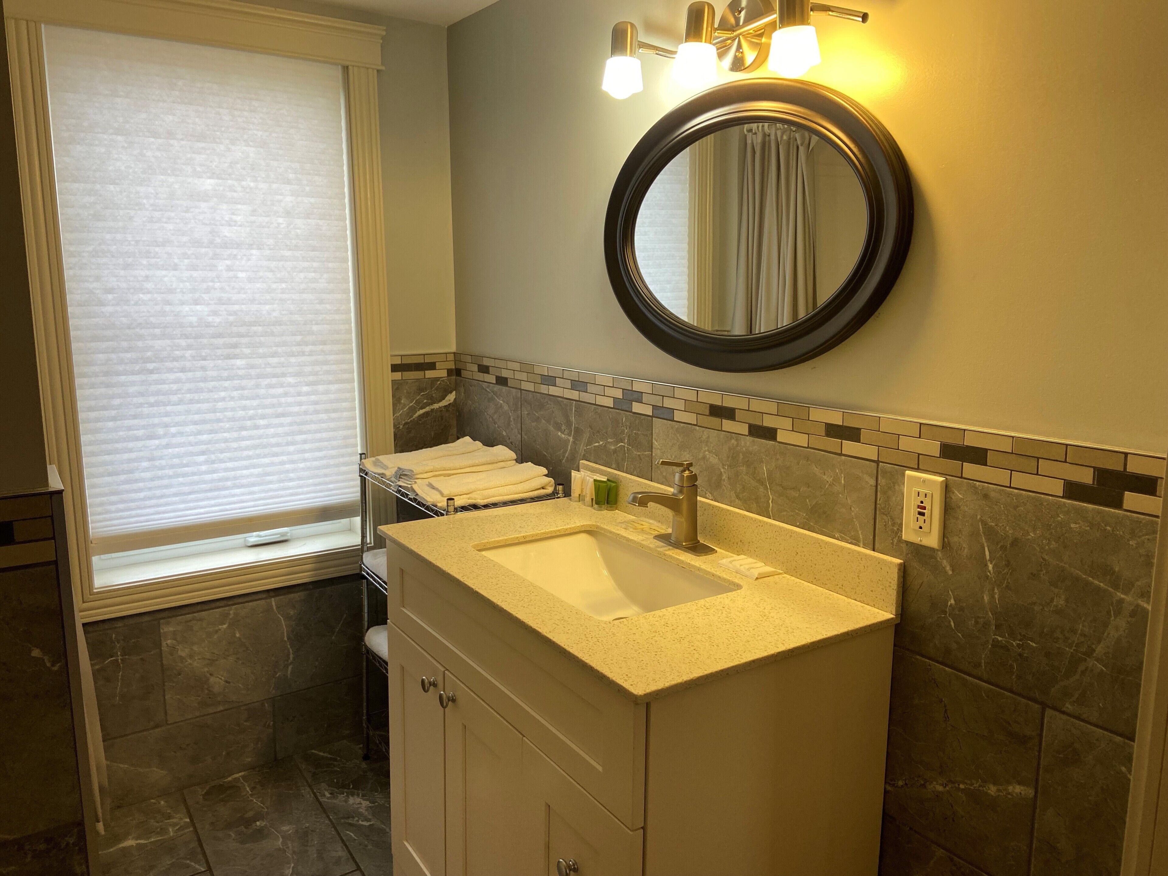 Une salle de bain équipée d’un lavabo, d’un miroir, d’un porte-serviettes et d’une fenêtre avec des stores.