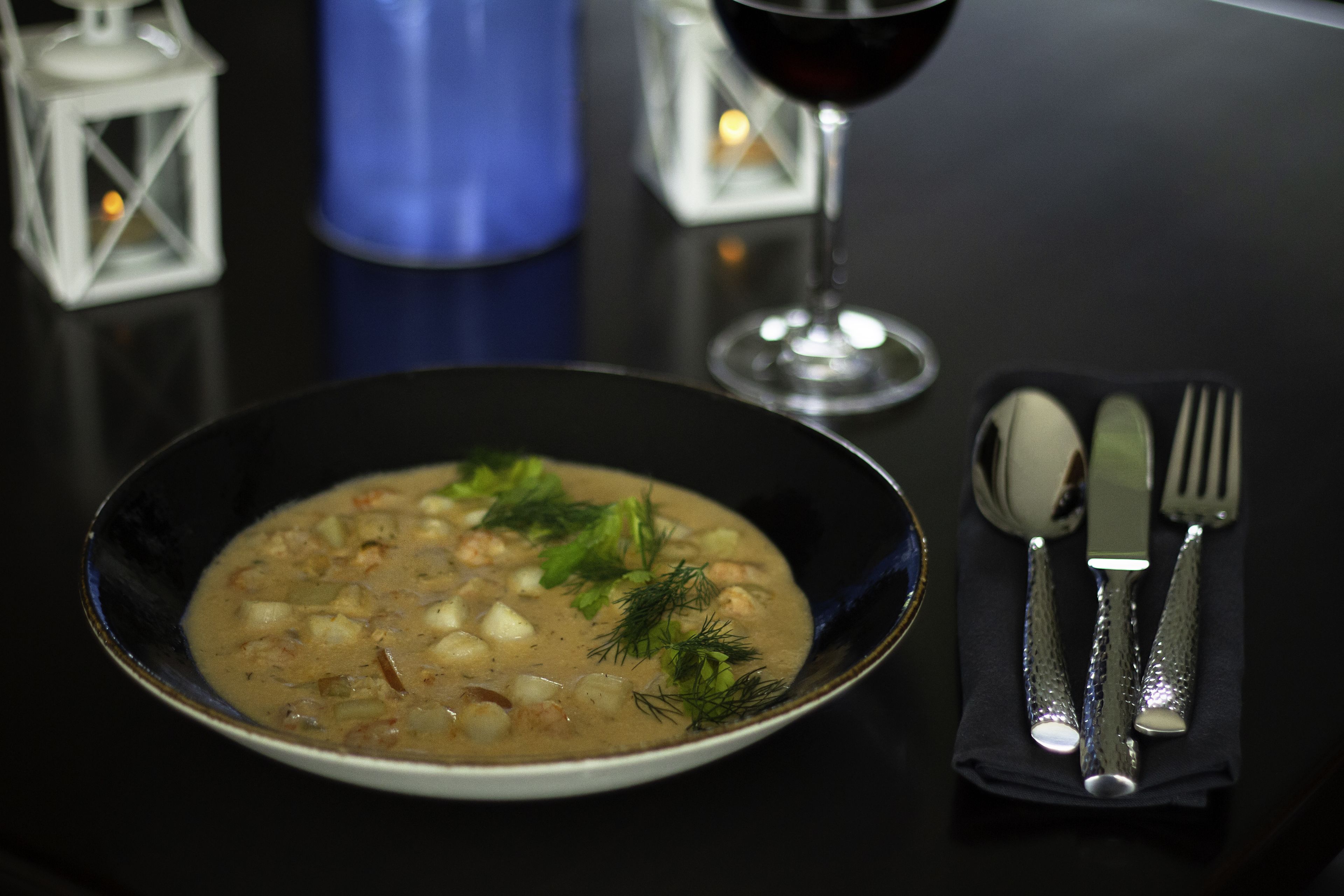 A bowl of soup with a creamy texture and garnished with herbs, accompanied by a glass of red wine, a blue candle, and cutlery on a black table.
