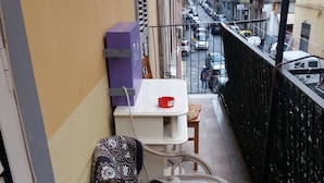 A narrow balcony with a wicker chair and a small table, overlooking a street with parked cars and buildings. - B&B Appartamenti Napoli (Naples)