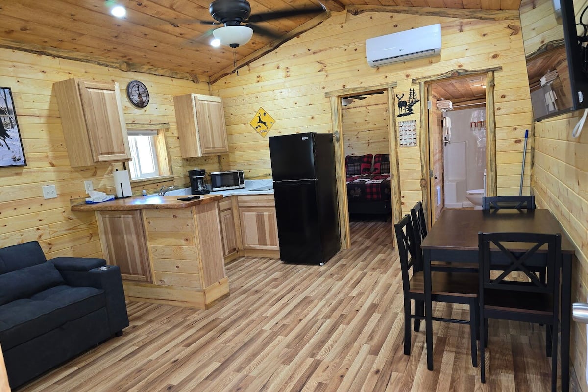A wooden cabin interior with a kitchen, dining area, and a black sofa.