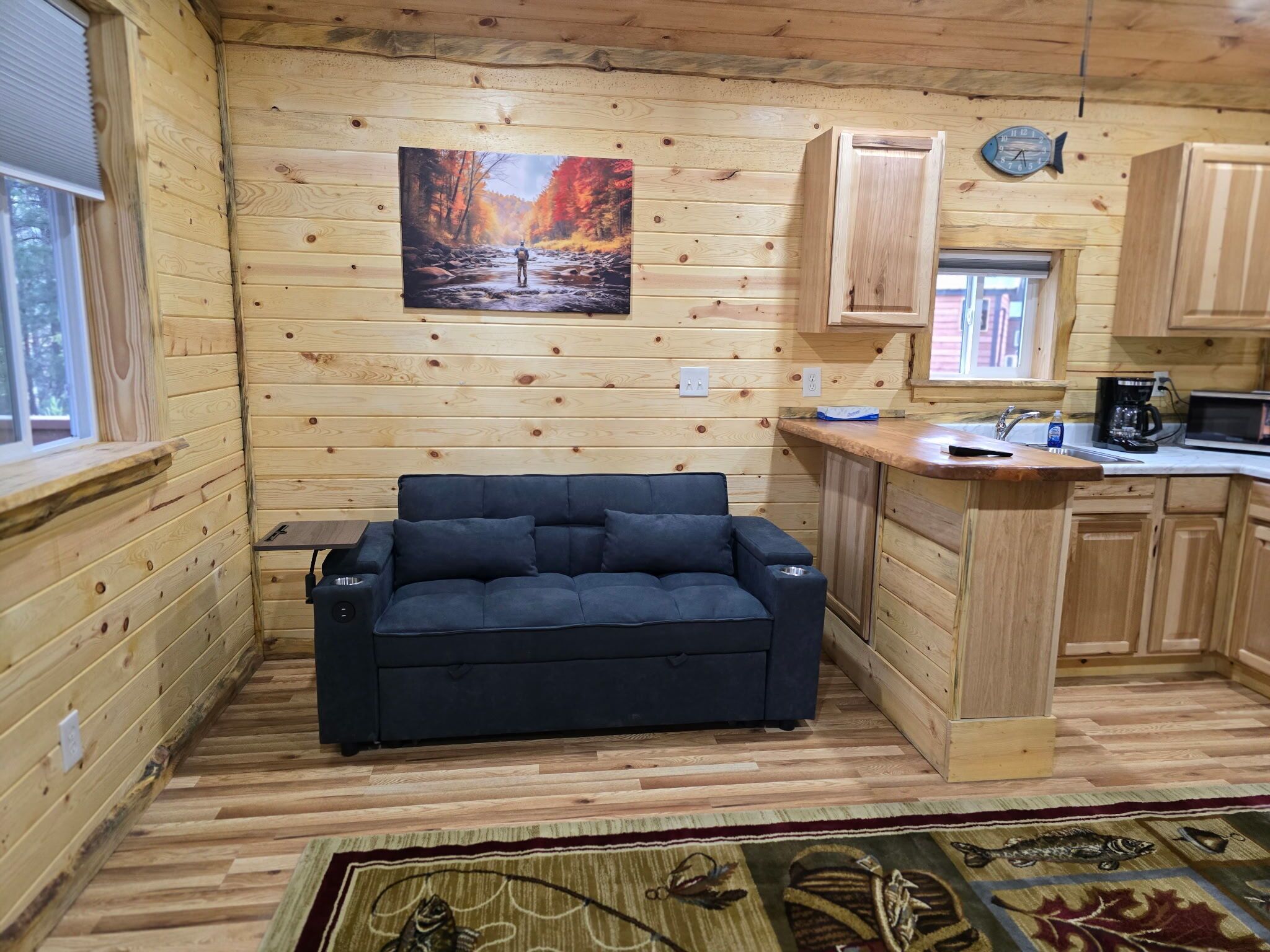 A cozy cabin interior with a sofa, wooden walls, a kitchen area, and a painting of a forest scene.