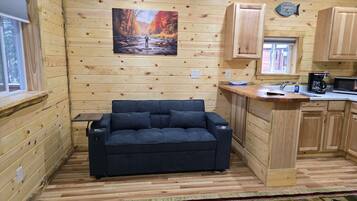 A cozy cabin interior with a sofa, wooden walls, a kitchen area, and a painting of a forest scene.