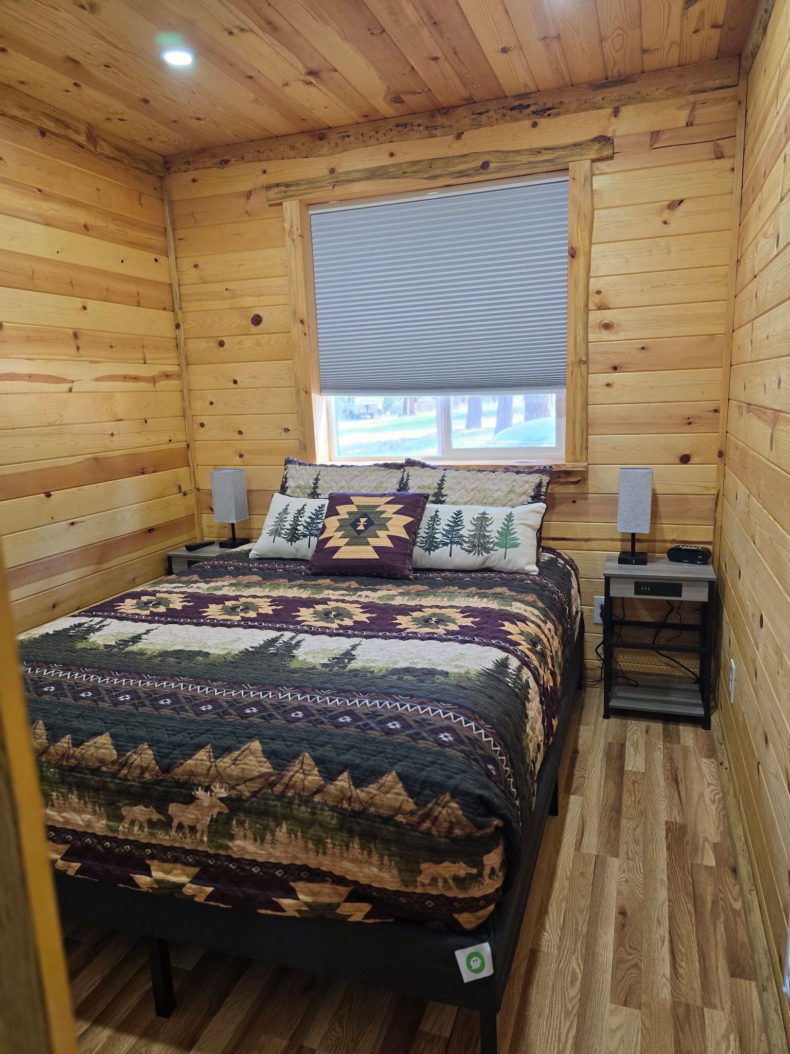 Ein Schlafzimmer in einer Holzhütte mit einem Bett, zwei Nachttischen, einem Fenster mit Jalousien und Blick auf ein Gebäude.