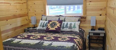 Ein Schlafzimmer in einer Holzhütte mit einem Bett, zwei Nachttischen, einem Fenster mit Jalousien und Blick auf ein Gebäude.