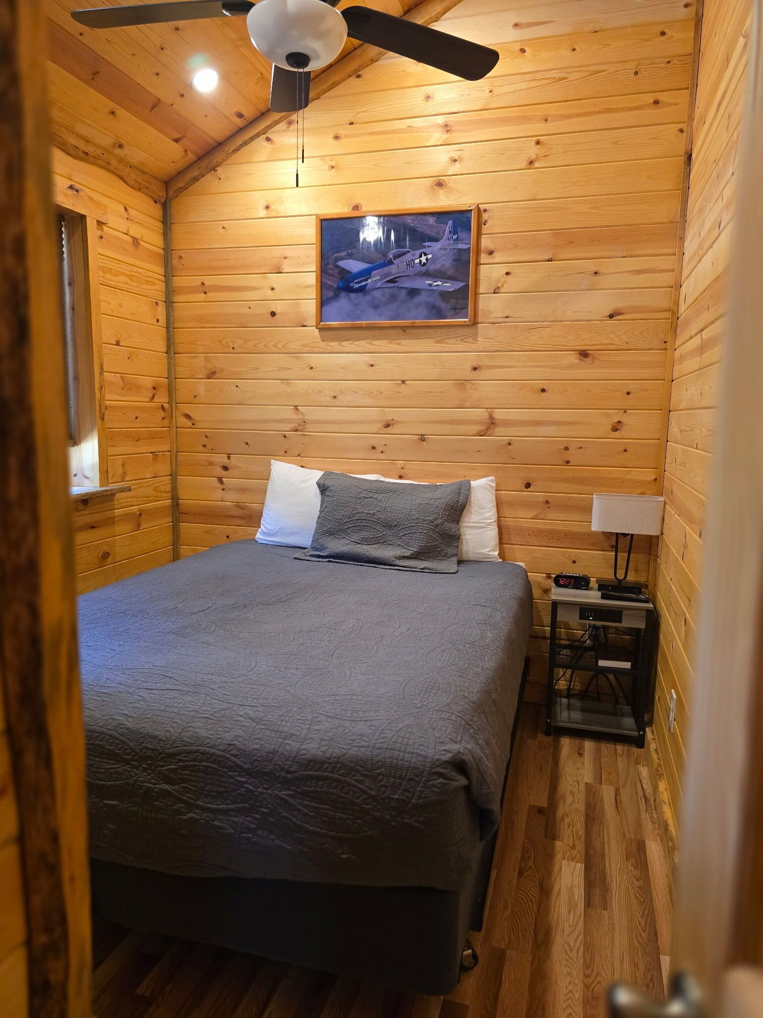 A cozy wooden cabin bedroom with a bed, pillows, a nightstand, a clock, and a ceiling fan.