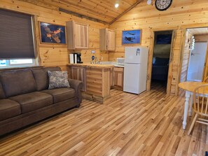 A cozy cabin interior with a wooden floor, a sofa, a kitchen area with a refrigerator, and a dining table. - Rimrock Lodge (Thompson Falls)