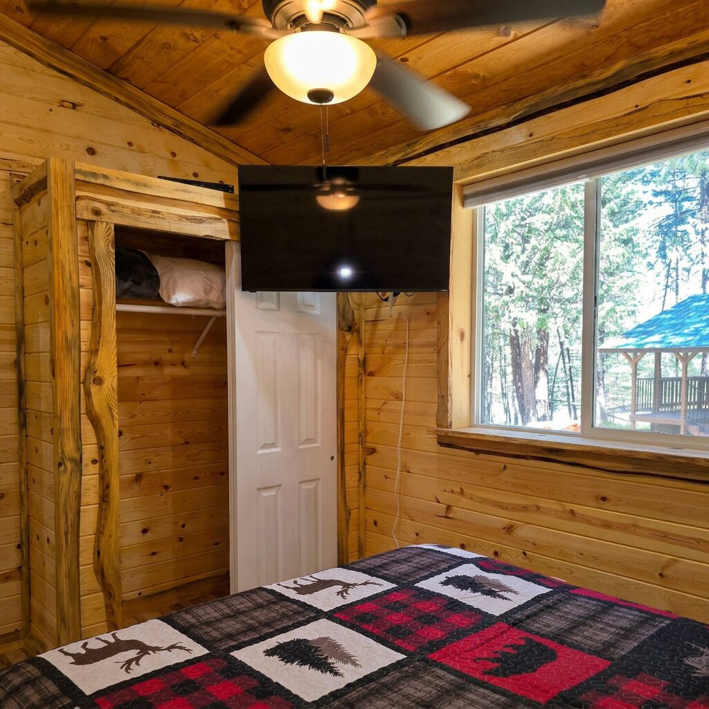 A wooden cabin room with a bed, a ceiling fan, a window with a view of trees, and a door.