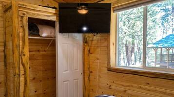 A wooden cabin room with a bed, a ceiling fan, a window with a view of trees, and a door.