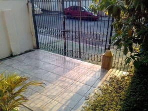 Un balcón con baldosas y una reja de metal, un coche rojo estacionado afuera y algo de follaje.