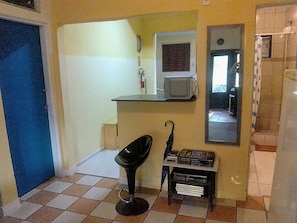 Una habitación pequeña con una puerta azul, una silla negra, una mesita con un libro, un espejo y un microondas.