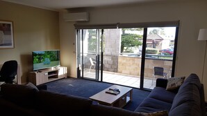 A living room with a TV, sofa, coffee table, and a sliding glass door leading to an outdoor area with a patio and a car parked outside. - Gillies st Apartments (Warrnambool)