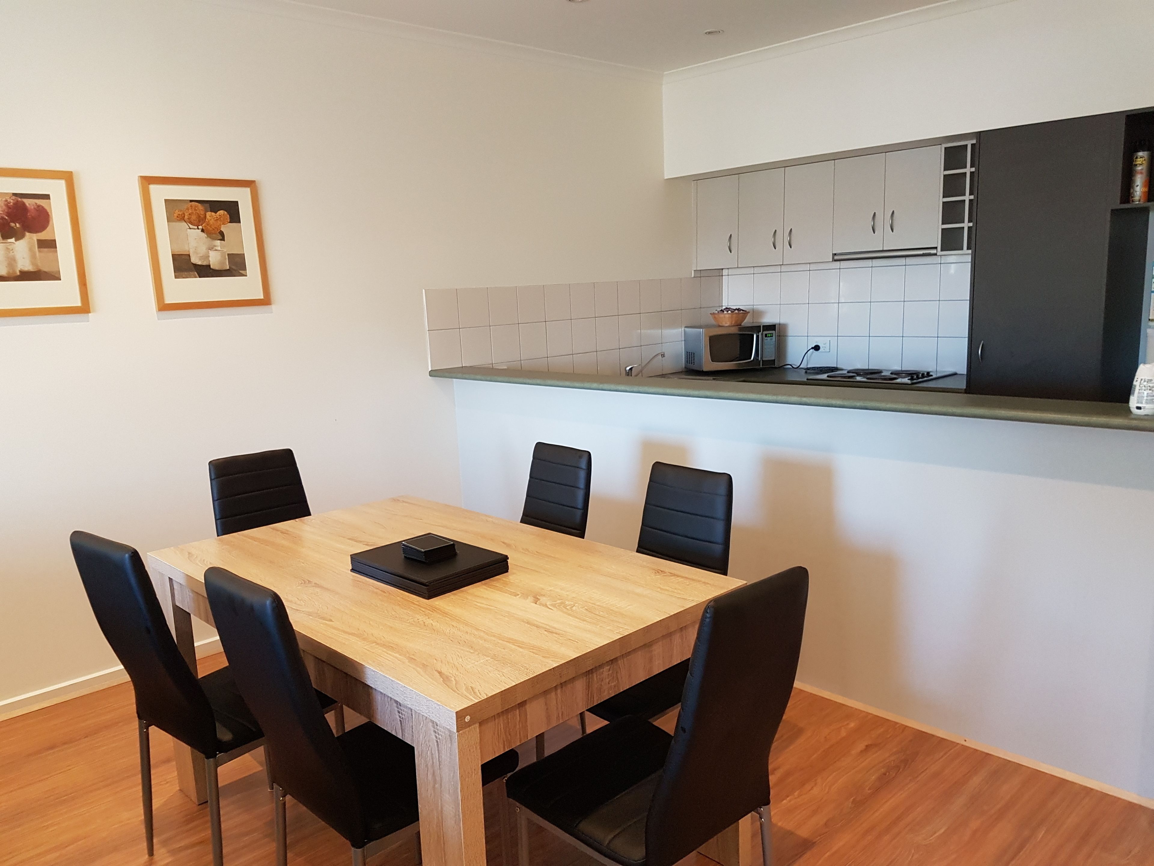 Ein Essbereich mit Holztisch und schwarzen Stühlen, eine Küche mit weißen Schränken und einer Mikrowelle sowie zwei gerahmte Bilder an der Wand.