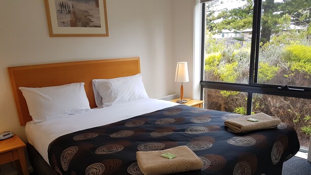 A hotel room with a large bed, a nightstand, a lamp, and a window with a view of greenery. - Gillies st Apartments (Warrnambool)