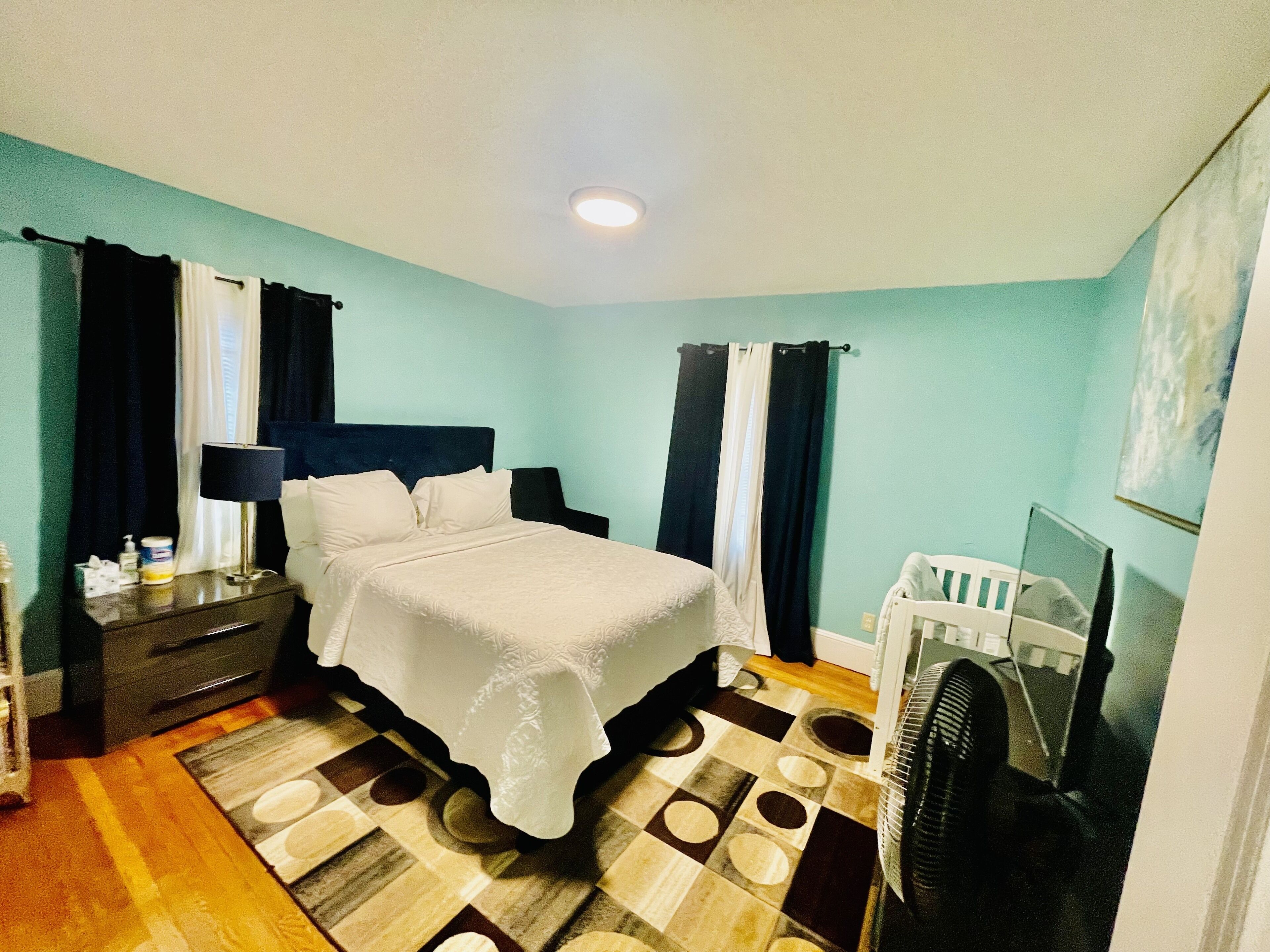 A hotel room with a bed, bedside table, lamp, curtains, a small desk, a chair, a fan, and a television.