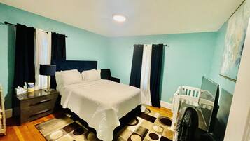 A hotel room with a bed, bedside table, lamp, curtains, a small desk, a chair, a fan, and a television.