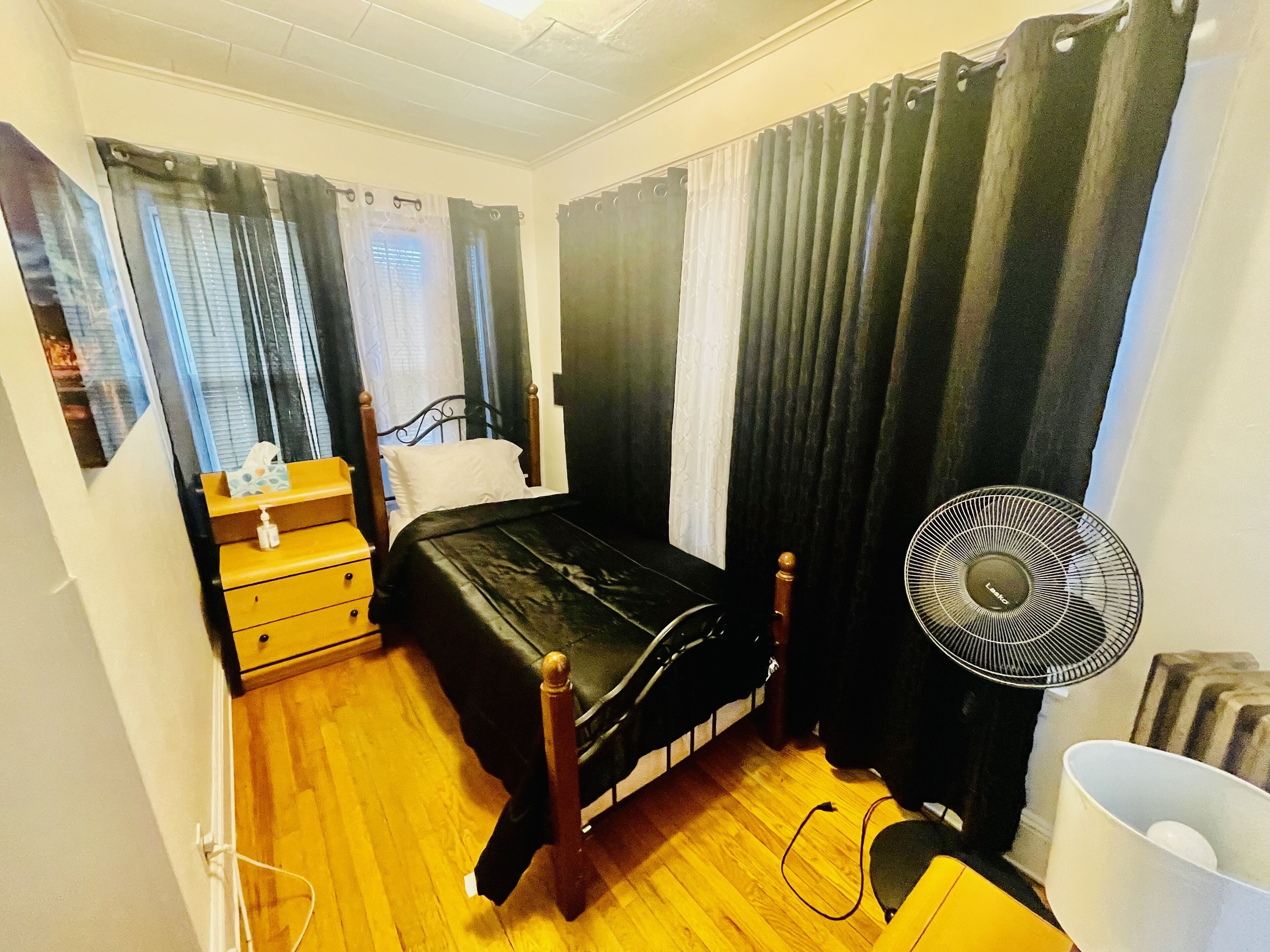Una camera da letto con un letto, un comodino con una lampada, una ventola e una finestra con tende.